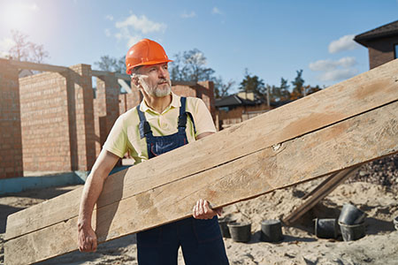 Kypäräpäinen rakennusalan työntekijä kantamassa lankkua rakennustyömaalla